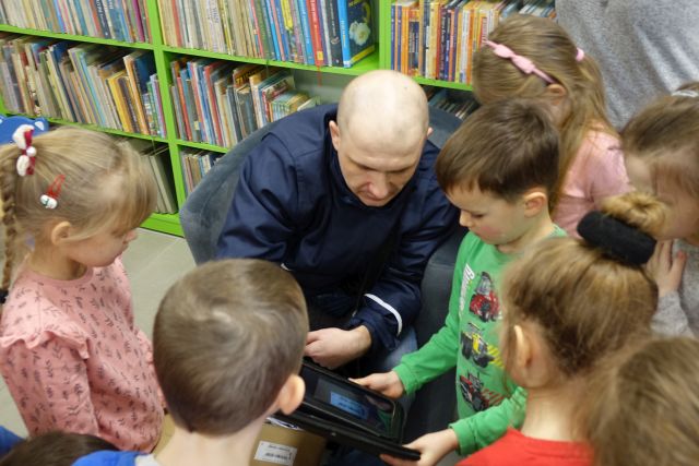 listonosz pokazuje dzieciom swoją torbę roboczą, w tle regały z książkami