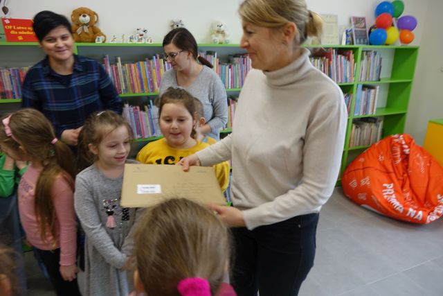 kobieta trzyma w rękach list w szarej kopercie, wokół niej stoi gromadka dzieci, w tle regały z książkami