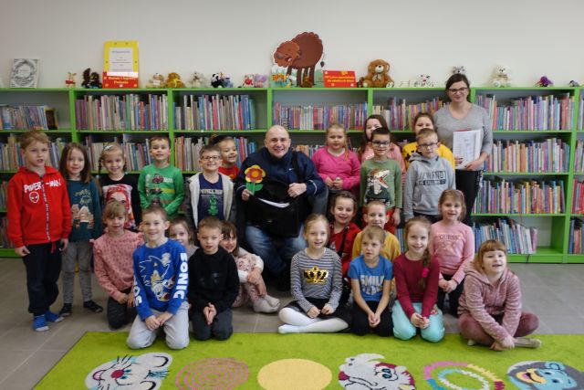 dzieci pozują do grupowego zdjęcia z listonoszem, w tle regały z książkami