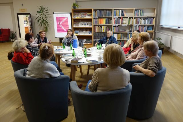 uczestnicy Dyskusyjnego Klubu Książki siedzą na siwych fotelach przy białych okrągłych stołach. W tle regały z książkami
