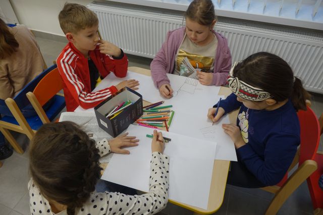 dzieci siedzą przy stolikach i rysują kredkami na kartkach koty