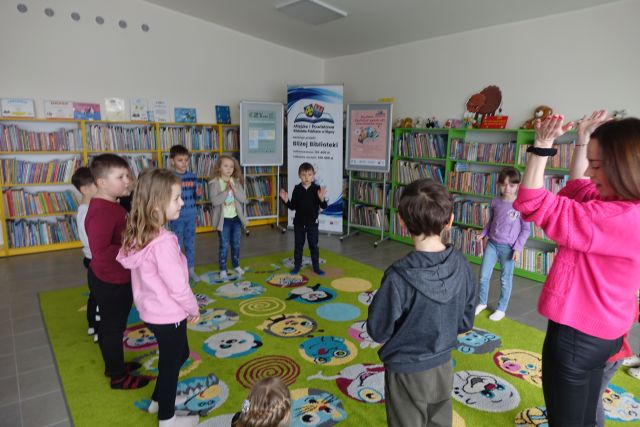 dzieci stoją na zielonym dywanie w postacie z bajek, w tle regały z książkami