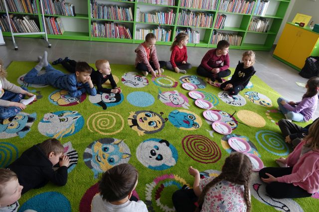 dzieci siedzą na zielonym dywanie przed nimi na papierowych talerzach wypisane po angielsku dni tygodnia, w tle regały z książkami