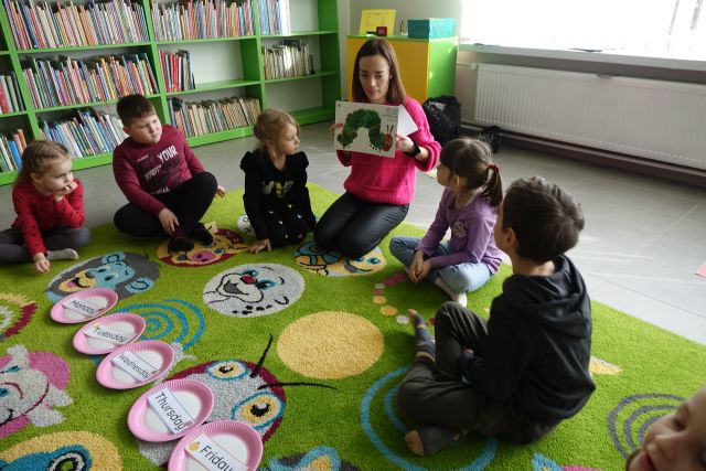 kobieta w różowej bluzce siedząca na zielonym dywanie pokazuje dzieciom siedzącym wokół niej na zielonym dywanie książkę z dżdżownicą na okładce, w tle okno, a z lewej strony regały z książkami