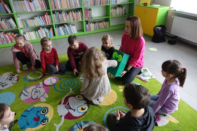 dzieci siedzą na zielonymdywanie, kobieta w różowej bluzce trzyma w dłoniach zielony karton, z którego dziewczynka wyciąga kartki