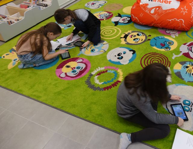 trzy dziewczynki siedzą na zielonym dywanie i za pomocą tabletów i zadaniem na kartce programują roboty