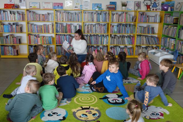 dzieci siedzą na dywanie, przed nimi na pufie siedzi kobieta w białym swetrze i czyta książkę, w tle kolorowe regały z książkami