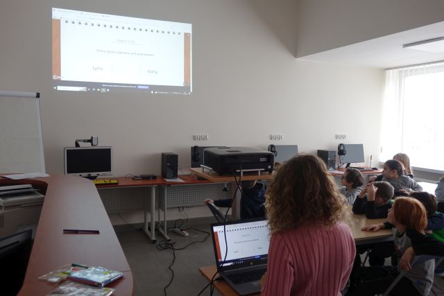 dzieci siedzą w sali komputerowej przy stołach, dziewczynka stoi przed laptopem i wyświetla prezentację, która wyświetla się na ekranie