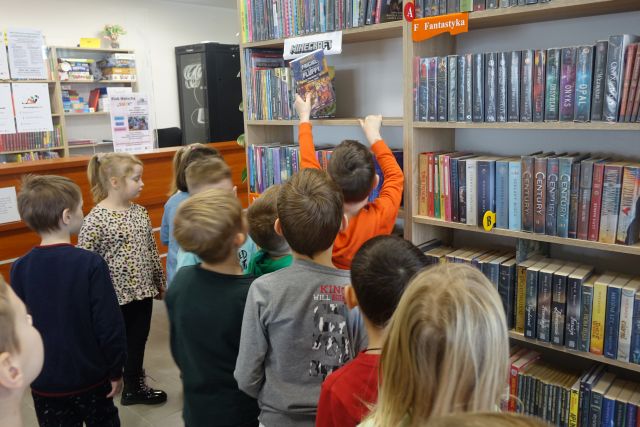 grupka dzieci stoi przed regałem z książkami, z lewej strony biurko