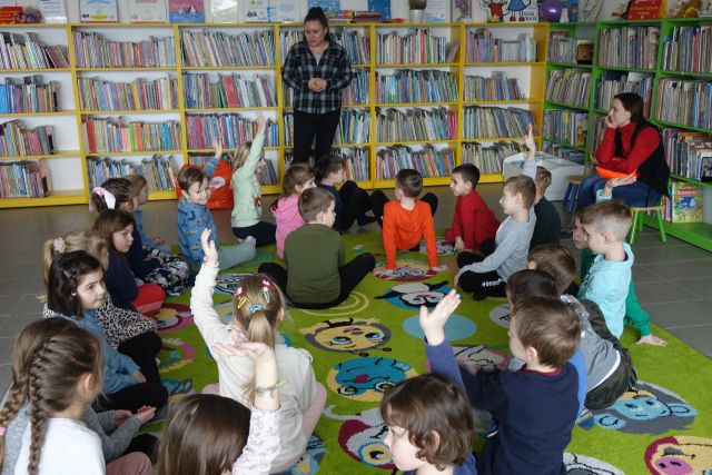 dzieci siedzą na zielonym dywanie, przed nimi stoi kobieta, w tle regały z książkami