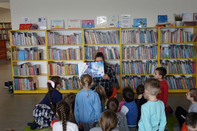 dzieci siedzą na dywanie przed nimi kobieta czytająca książkę, w tle regały z książkami