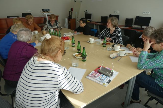 seniorki siedzą przy połączonych stołach i piszą na kartkach, przed nimi filiżanki z kawą, ciastka i woda w butelkach
