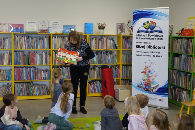 kobieta pokazuje dzieciom siedzącym na kolorowym dywanie książkę, w tle regały z książkami