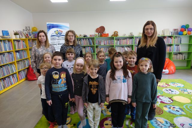 dzieci i trzy kobiety pozują do zdjęcia, w tle kolorowe regały z książkami i plansza z informacja o projekcie Bliżej Biblioteki i logo biblioteki