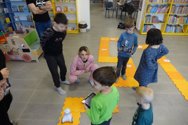 dzieci za pomocą tabletów sterują robotami, w tle regały z książkami
