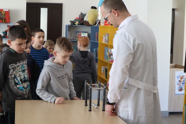 mężczyzna w białym faruchu pokazuje doświadzcenie z magnesami, w kolejce stoją dzieci chcące spróbować, w tle regały z książkami