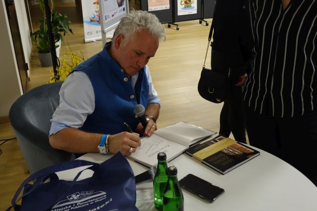 mężczyzna w niebieskim bezrękawniku i białej koszuli podpisuje książkę, przed nim stoi kobieta ubrana na czarno