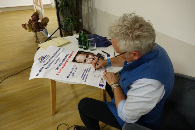 mężczyzna w niebieskim bezrękawniku i białęj koszuli w okularach podpisuje się na plakacie zapraszającym na spotkanie