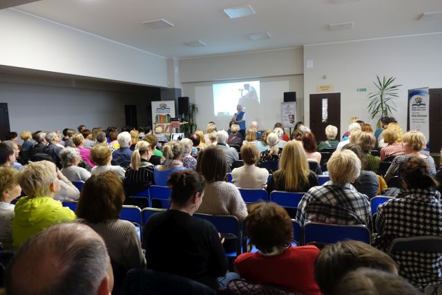 tyłem siedzi około 150 uczestnikó spotkania z Jarosłąwem Kotem, przed nimi prezentacja na ekranie stojący imężczyzna w niebieskim bezrękawniku
