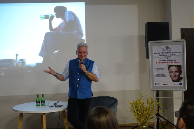 mężczyzna w niebieskim bezrękawniku i białęj koszuli trzyma w ręce mikrofon, w tle wyświetlana prezentacja