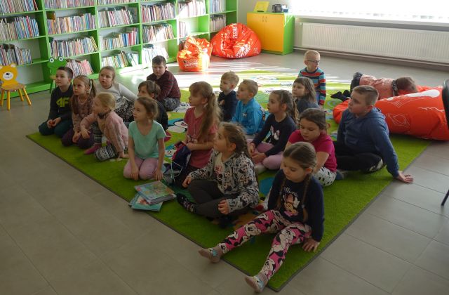 grupa dzieci siedzi na zielonym dywanie, w tle regały z książkami