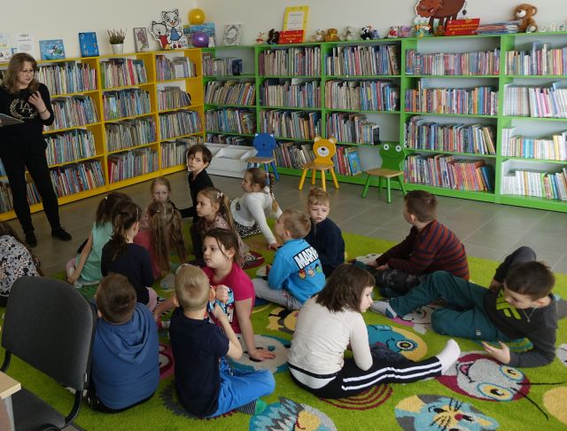 dzieci siedzą na zielonym dywanie, przed nimi stoi kobieta z książką ubrana na czarno, w tle regały z książkami