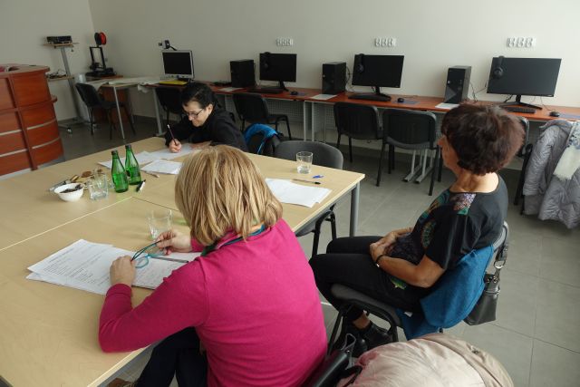 trzy seniorki siedzą przy stołach i piszą na kartkach, przed nimi flipchart i biurko, z prawej strony biurka z komputerami