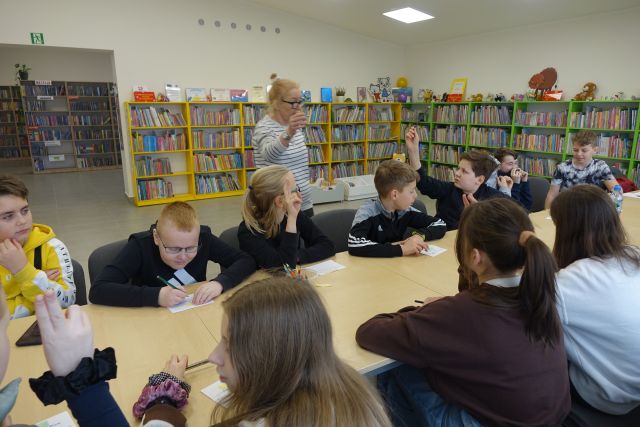 młodzież siedząca przy stołąch pisze na kartkach, za nimi kobieta, w tle regały z książkami