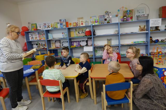 dzieci siedzą przy stolikach i słuchają opowiadającej kobiety, w tle kolorowe regały