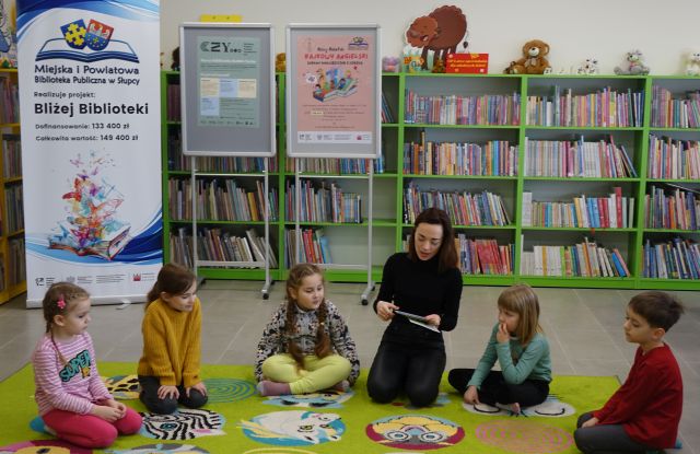 kobieta ubrana na czarno siedzi z dziećmi na zielonym dywanie, w tle plakat z nazwią projektu i logo biblioteki oraz regały z książkami