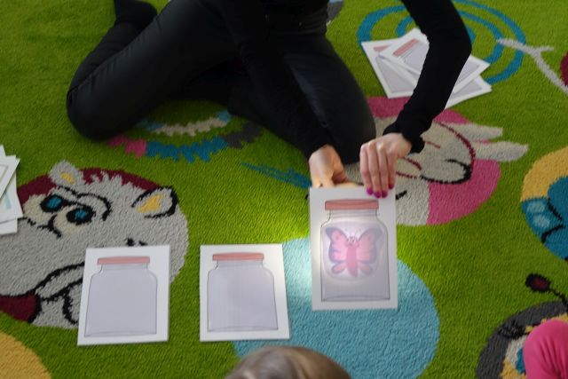 kobieta ubrana na czarno pokazuje dzieciom podświetlone kartki ze słoikami