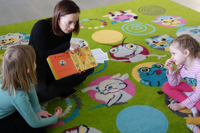 kobieta siedząca na zielonym dywanie pokazuje dzieciom książkę