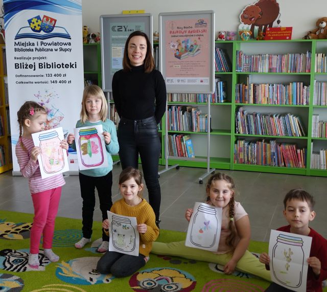 kobieta ubrana na czarno i dzieci z ilustracjami wykonanymi przez siebie przedstawiającymi robaki w słoikach pozują do zdjęcia