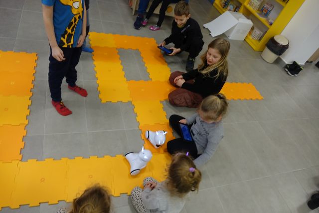 dzieci siedzą na podłodze, przed nimi po pomarańczowych puzlach jeżdżą roboty Photon