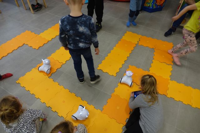 dzieci siedzą na podłodze i stoją, przed nimi po pomarańczowych puzlach jeżdżą roboty Photon