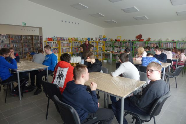 uczestnicy warsztatów siedzą przy stolikach, przed nimi stoi kobieta, w tle regały z książkami