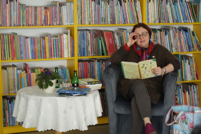 Kobieta siedzi na siwym fotelu i czyta książkę, obok niej stoi przykryty beżowym obrusem stolik, w tle regały z książkami