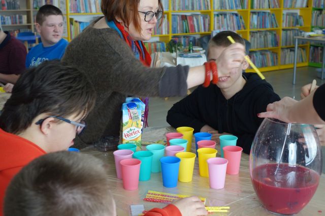młodzi ludzie siedzą przy stolikacj, przed nimi kobieta stoi i miesza sok, na stole stoją kolorowe kubki