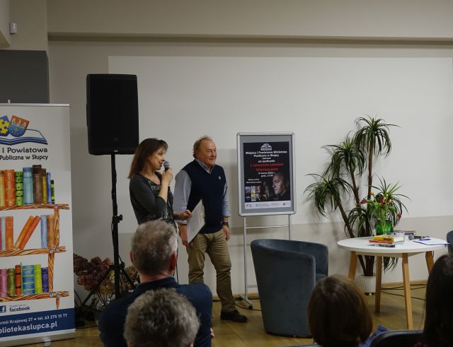 kobieta z mikrofonem ubrana na ciemno stoi obok mężczyzny w jasnej koszuli i granatowym bezrękawniku, w tle kolumna i plakat zapraszający na spotkanie