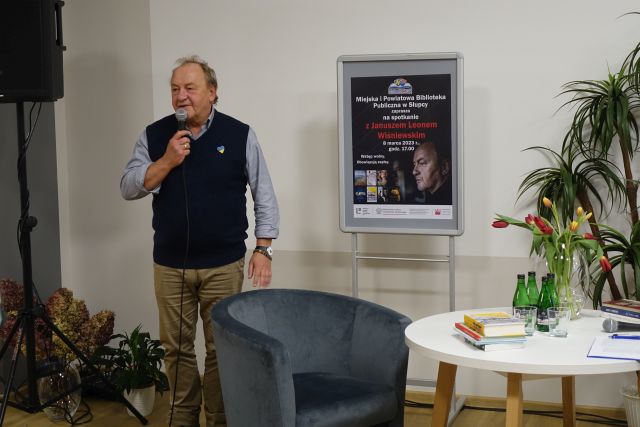 mężczyzna w granatowym bezrękawniku stoi z mikrofonem, w tle plakat zapraszający na spotkanie, obok siwy fotel