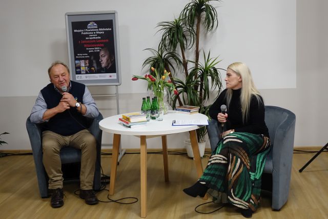 mężczyzna w granatowej kamizelce i kobieta w czarnym swetrze siedzą przy białym stole okrągłym i trzymają w dłoniach mikrofony, w tle duża sztuczna palma i plakat zapraszający na spotkanie