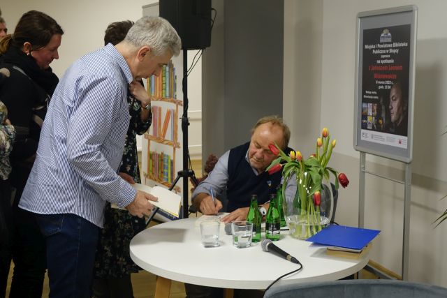 mężczyzna w granatowej kamizelce podpisuje siedząc przy białym okrągłym stole książkę, przed nim stoi mężczyzna z książkką