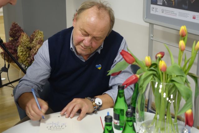 mężczyzna w granatowej kamizelce podpisuje książkę siedząc przy białym okrągłym stoliku, na któym stoją w wazonie żółte i czerwone tulipany