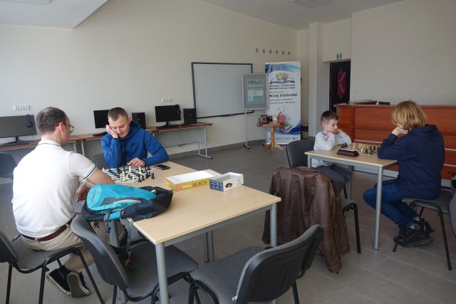 3 chłopców i mężczyzna grają w szachy siedząc przy stolikach, w tle sala komputerowa