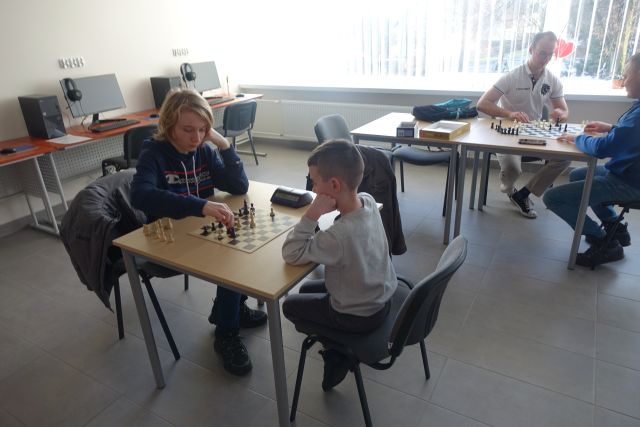 3 chłopcó i mężczyzna siedzą przy stolikach i grają w szachy, w tle sala komputerowa