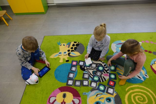 dzieci siedzące na zielonym dywanie kodują przy użycie tabletu trasę robota