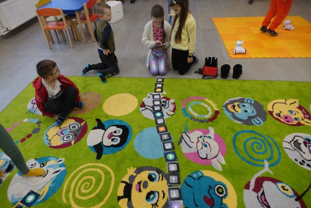 dzieci siedzące na zielonym dywanie kodują przy użycie tabletu trasę robota
