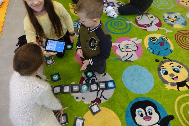 dzieci siedzące na zielonym dywanie kodują przy użycie tabletu trasę robota