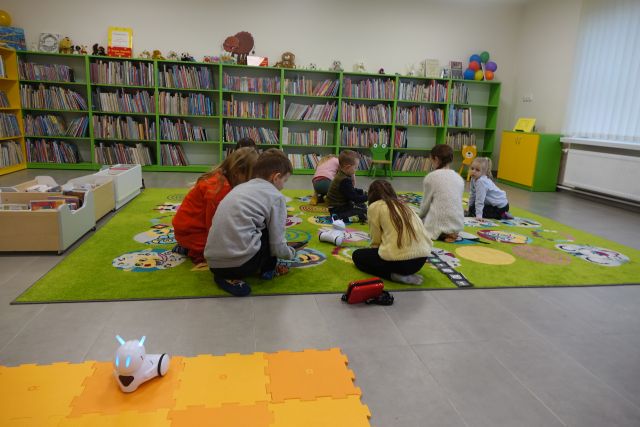 dzieci siedzą na zielonym dywanie i sterują przy pomocy tabletów robotami, w tle regały z książkami