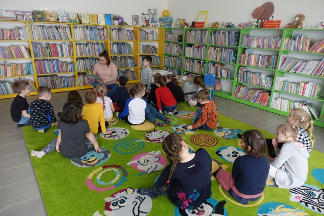 dzieci siedzą na zielonym dywanie, przed nimi kobieta z książką, w tle regały z książkami